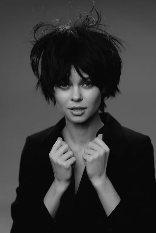 Black and white portrait of a young woman with messy hair wearing a black blazer, expressing confidence and intensity.