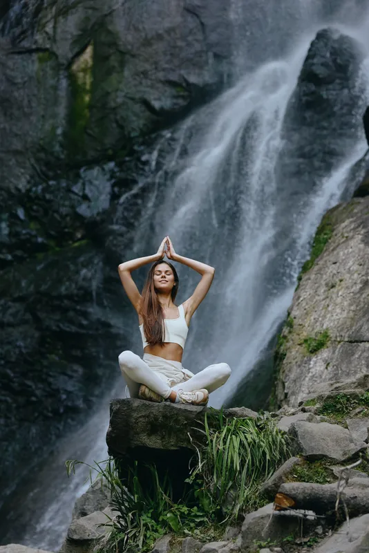 woman meditating outdoors near waterfall in white activewear sits cross-legged on rock with calm expression surrounded by lush greenery in peaceful nature setting