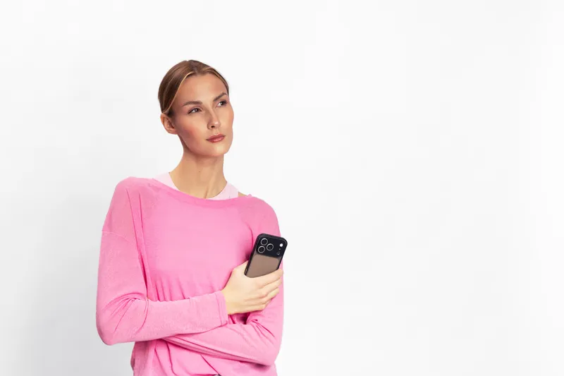 Young woman in pink sweater holds smartphone and looks sideways with thoughtful expression on white background. Concept for communication, technology, and contemplation.