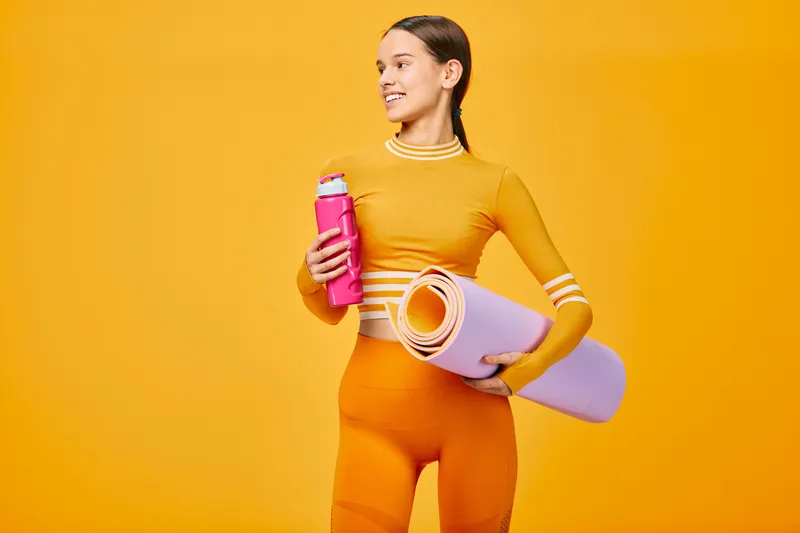 Woman in a bright fitness outfit holds a water bottle and rolled yoga mat against a vibrant yellow backdrop, ready for a workout or fitness session.