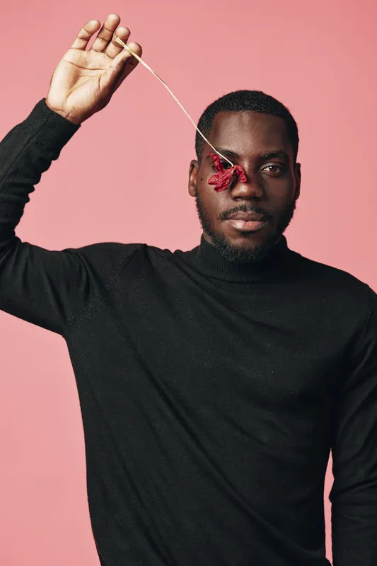 African man with dark skin wearing black sweater holding red flower near eye on pink background in creative conceptual visual style portrait with artistic expression and fashion pose