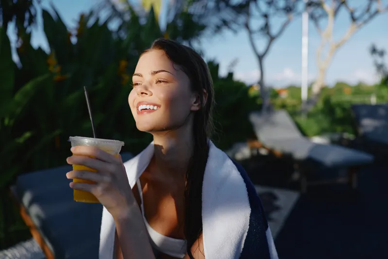 Cheerful woman outdoors, holding a cold beverage, smiling with eyes closed, warm sunlight, relaxed posture, tropical plants in the background, vibrant natural colors, casual summer moment, with soft