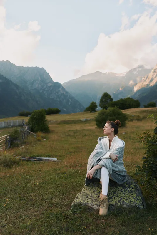 Young woman wrapped in a cozy shawl sits on a rock in a mountain meadow enjoying the peaceful natural landscape during a calm afternoon.