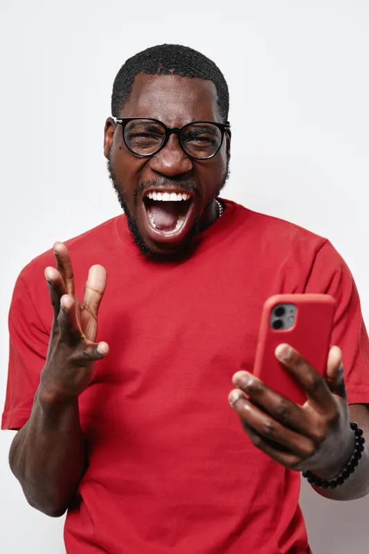 African American man with dark skin wearing glasses and red shirt shouting with excitement while holding smartphone in hand on white background.