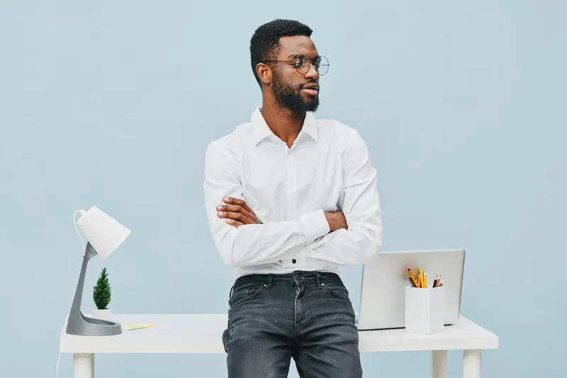 Confident young man with glasses standing in modern office environment, dressed in white shirt, crossing arms, looking away thoughtfully, minimalistic workspace with laptop and desk accessories, profe