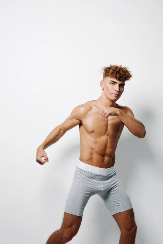 Muscular young man in light grey shorts with defined abs posing with arms raised on white background. Athletic body, youthful fitness, curly hair and confident expression, isolated studio portrait.