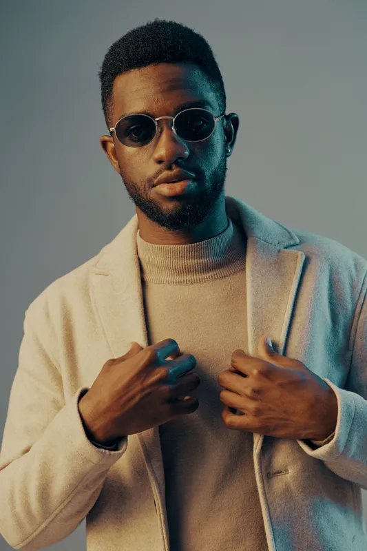 Stylish young black man in sunglasses wearing a beige coat and sweater against a soft blue background, exuding confidence and modern fashion