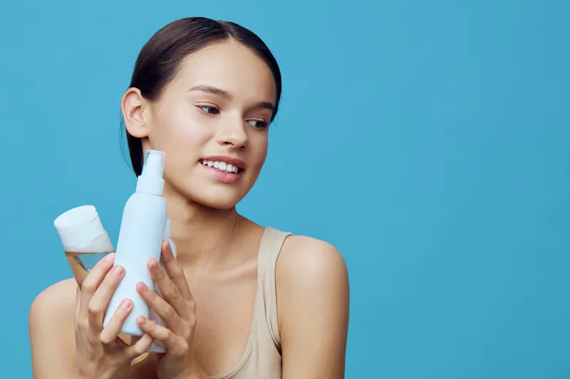 Young model showcasing skincare products on blue background, smiling and conveying confidence with beauty essentials, healthy skin, and self care focus