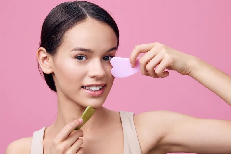 Young woman with smooth skin holding skincare tools, smiling brightly against a pink background, representing beauty and self care routines