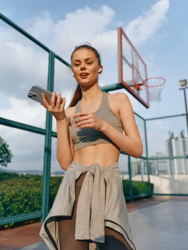 Спортивная девушка-модель в спортивном топе и леггинсах стоит на открытой баскетбольной площадке, используя телефон, на заднем плане виден городской пейзаж