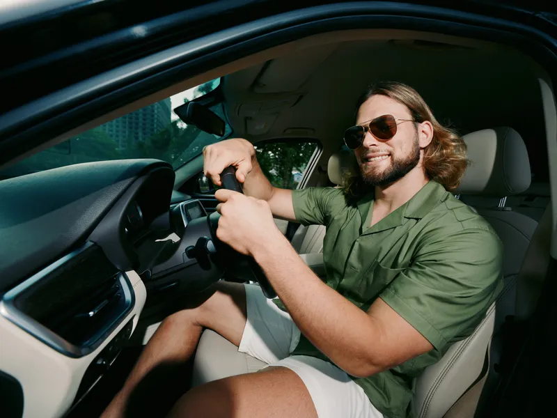 Smiling man wearing sunglasses drives a car, gripping the steering wheel with focus and energy. A sunlit interior creates a casual modern road trip vibe.