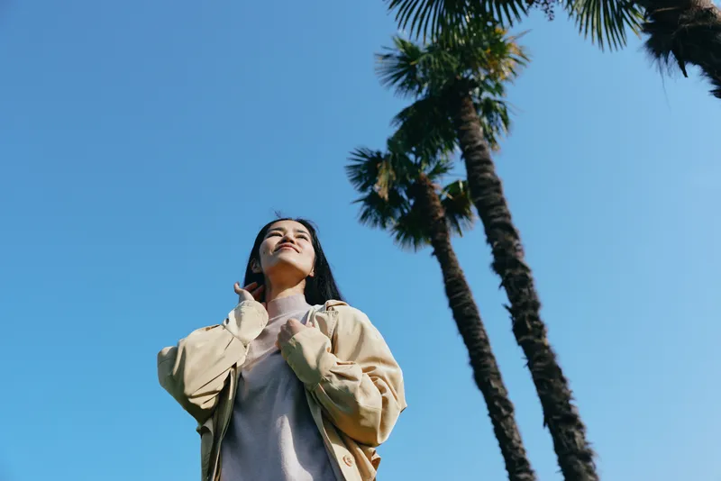 Outdoor scene with a woman looking up at a bright blue sky framed by tall palm trees, wearing a light jacket, soft sunlight, and a serene, hopeful mood during golden hour.