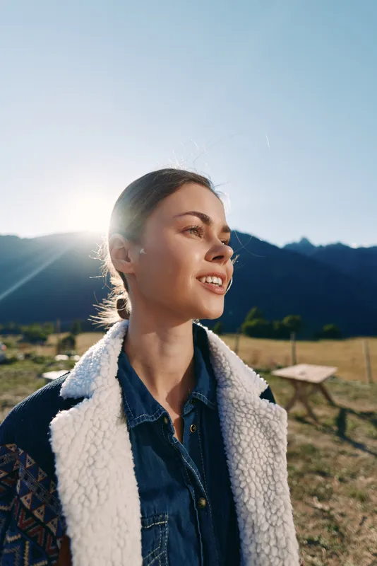 woman portrait mountains sunlight smile countryside, young female smiling in denim and shearling jacket, outdoor lifestyle backlit at golden hour, serene rural scenery and relaxed expression