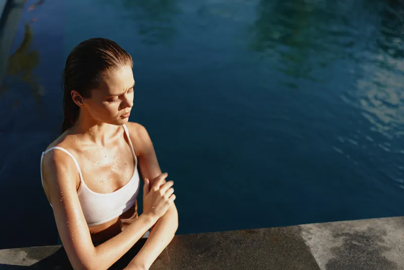 Young woman in white sportswear sitting by the pool, drying her arm with sunlight on her face, enjoying outdoor fitness and healthy lifestyle near water surface.