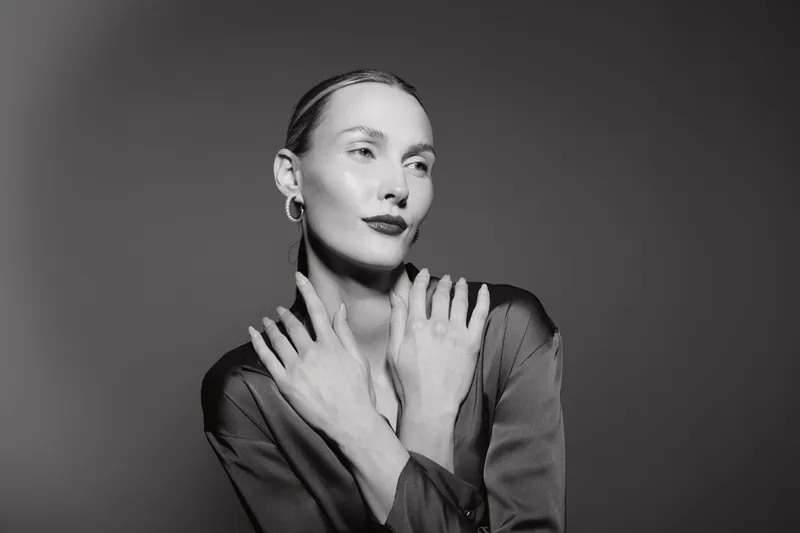 Elegant white woman with pale skin posing in a studio with crossed hands, wearing hoop earrings and a dark satin blouse.