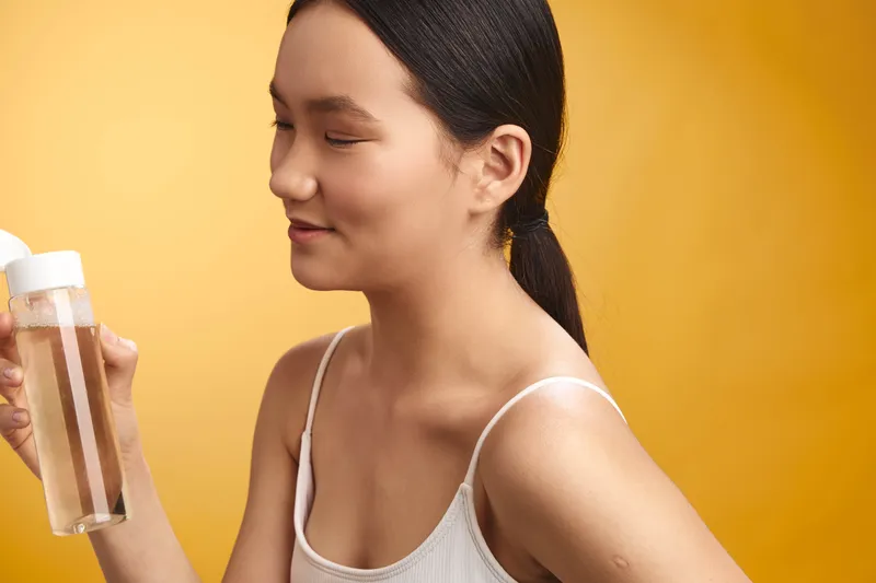 asian woman holds a bottle against a warm orange backdrop, focused on the container. she wears a light tank top, creating a calm, simple health or skincare moment.