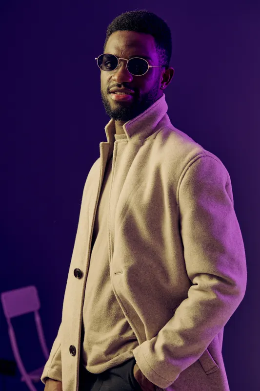 Stylish man in a beige coat and sunglasses exuding confidence against a purple background, showcasing modern fashion trends and urban vibes