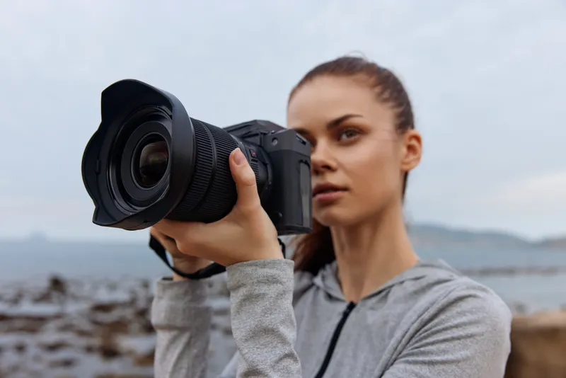 Young woman capturing a moment with a camera by the sea, wearing a sporty outfit in a serene setting, highlighting passion for photography and nature