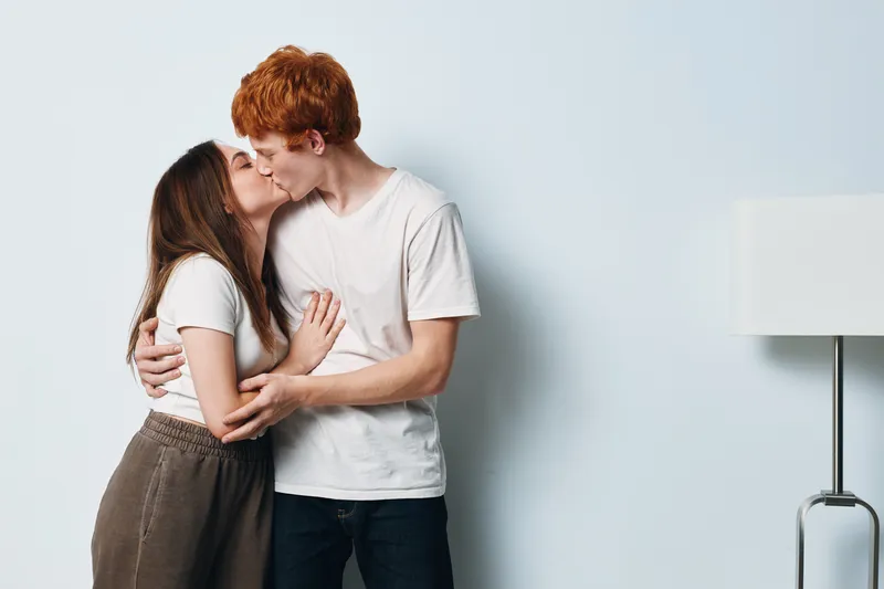 Couple kissing and embracing in casual clothing standing in minimalistic room with lamp and light background. Young man and woman in affectionate moment of love and connection.
