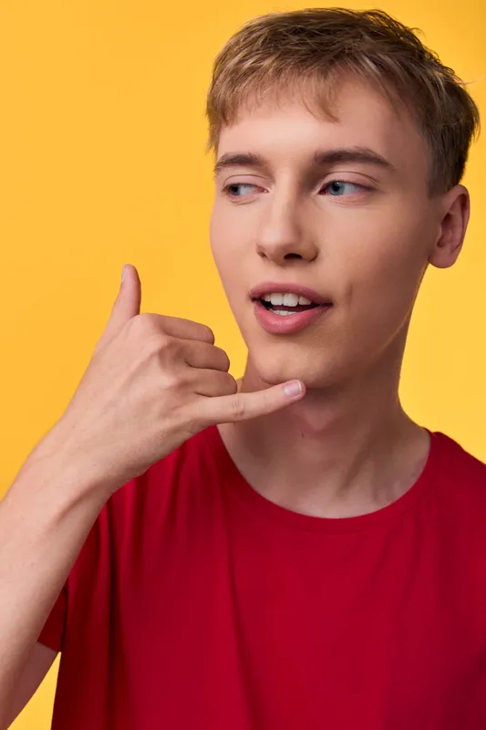 Hand gesture indicating call, young man in red shirt against a yellow background, casual lifestyle, friendly expression, communication cue, modern fashion and positive vibe