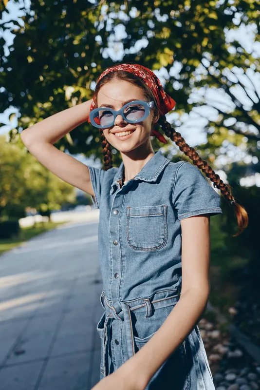 smiling woman in denim dress wearing blue sunglasses and red bandana outdoors on sunny day with green trees background