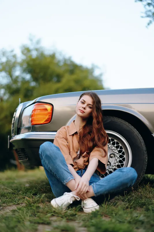Young woman sitting next to a vintage car, wearing casual attire and sneakers, conveying a relaxed mood in nature with green background and soft light.