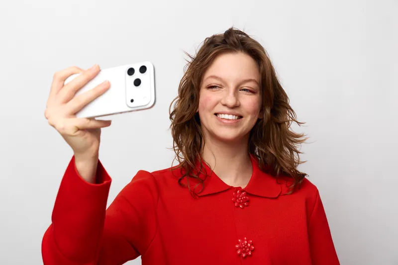 Smiling woman in red sweater takes selfie with white smartphone on white background. Casual lifestyle, technology use, social media concept with happy young adult female portrait.