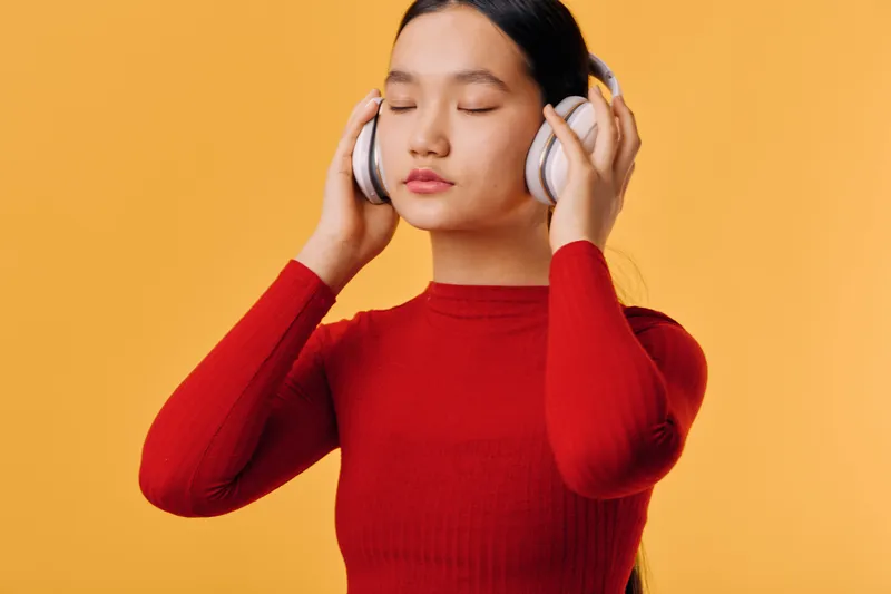 Woman with closed eyes wearing headphones and red sweater listening to music on orange background. Relaxed female enjoying audio entertainment with peaceful expression and casual style.
