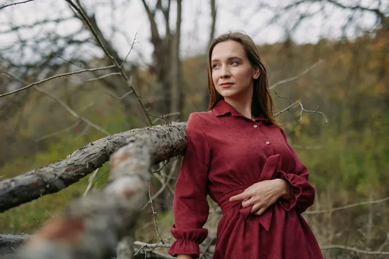 A candid portrait of a woman in a burgundy dress beside a weathered log in a quiet woodland, conveying authenticity through natural light and subtle emotion.