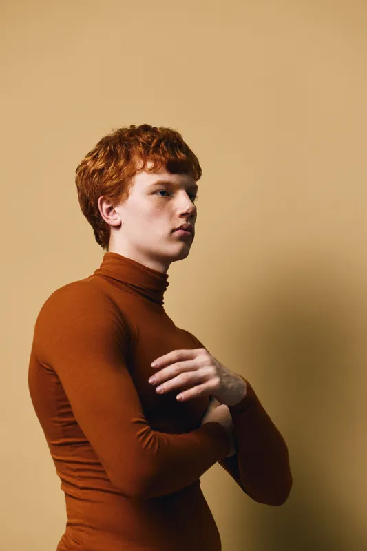 Young man with curly red hair wearing brown turtleneck posing in profile on beige background studio portrait of confident male model with folded arms and serious expression.