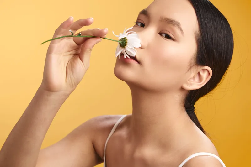 asian woman holds a delicate flower near her lips in a bright studio, creating a fresh, relaxed mood with soft light and warm color that highlights natural beauty.