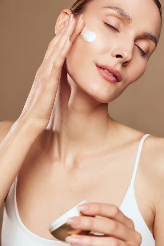 A young woman with clear skin applies moisturizer cream on her cheek with closed eyes. The natural beauty concept shows skincare routine and self care in a bright studio setting.