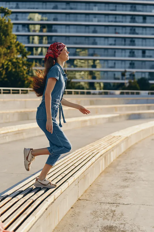 lifestyle woman in modern boho-western denim street style walks outdoors with a red bandana and casual sneakers under warm natural light, film color effect