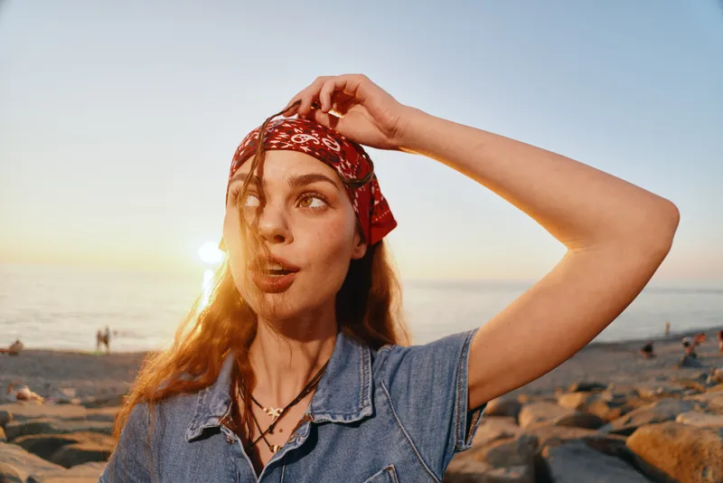 young woman wearing red bandana and denim shirt at sunset beach, looking thoughtful with sea and rocks background in warm golden light