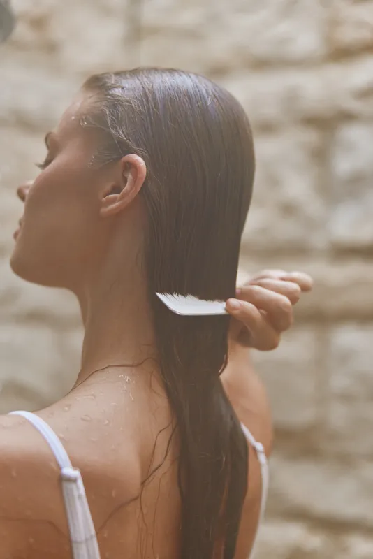 Woman combing wet hair with white comb near textured beige wall, natural light highlighting smooth skin and relaxed mood in warm neutral tones.
