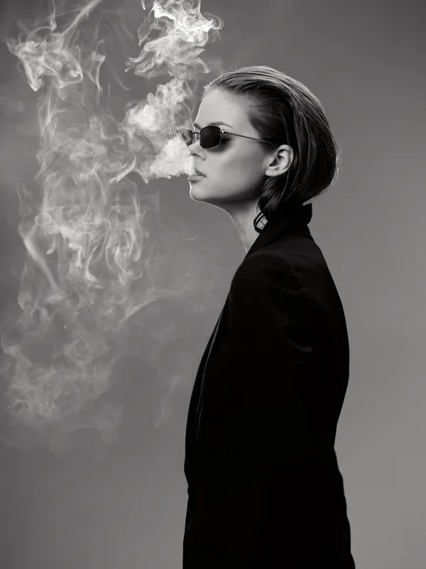 Black and white portrait of a woman wearing sunglasses exhaling smoke in a dramatic pose.