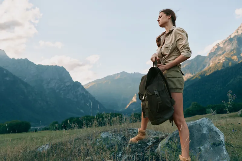 Woman hiking outdoors with a backpack, standing on rocky ground, sunlit mountains and valley scenery, casual athletic mood, natural colors and breeze