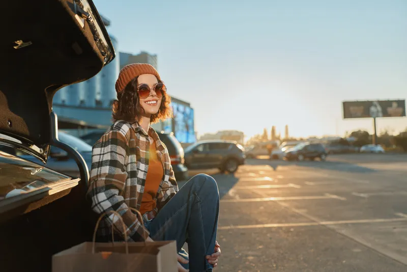 Woman shopping with bags by car trunk at parking lot during sunset, smile wearing sunglasses and beanie, candid lifestyle portrait with golden hour glow, authenticity and emotional storytelling.