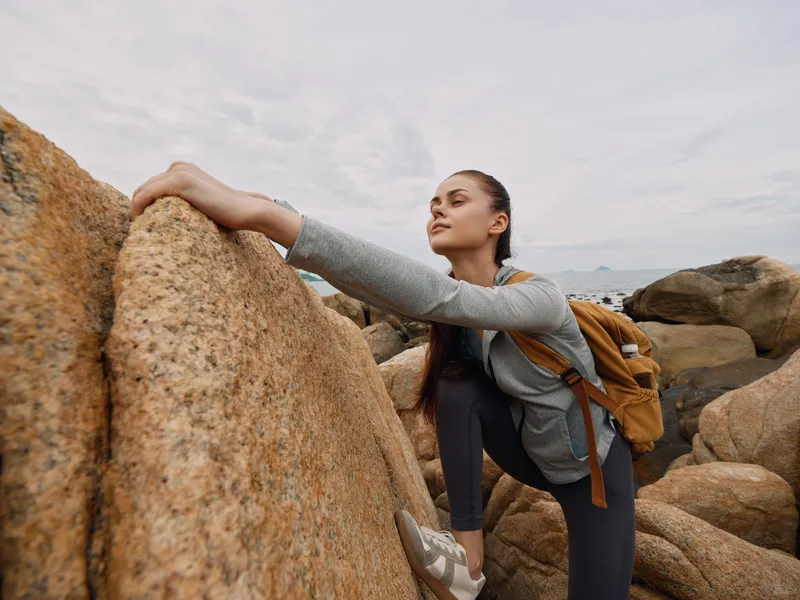 Active young woman climbing rocks in outdoor adventure, wearing casual sport outfit and backpack, showcasing determination and focus in natural landscape