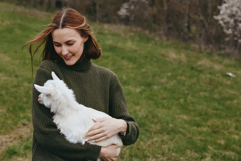 Woman in a chunky knitted sweater hugs a white goat kid in a grassy field. Natural light, candid lifestyle moment, warm mood, gentle affection between human and animal in an outdoor nature scene.