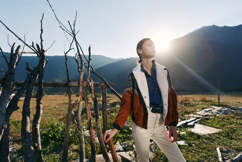 Woman in patterned jacket standing in a field with mountains and warm sunlight, posing near rustic wooden sticks and rocks, fashion portrait and lifestyle in natural outdoor landscape.