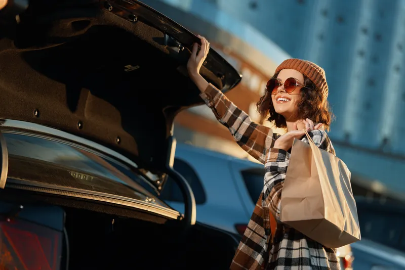 Woman shopping by car trunk with smile and bag in urban sunlight, candid lifestyle moment with golden hour glow, authenticity and emotional storytelling of mindful living.