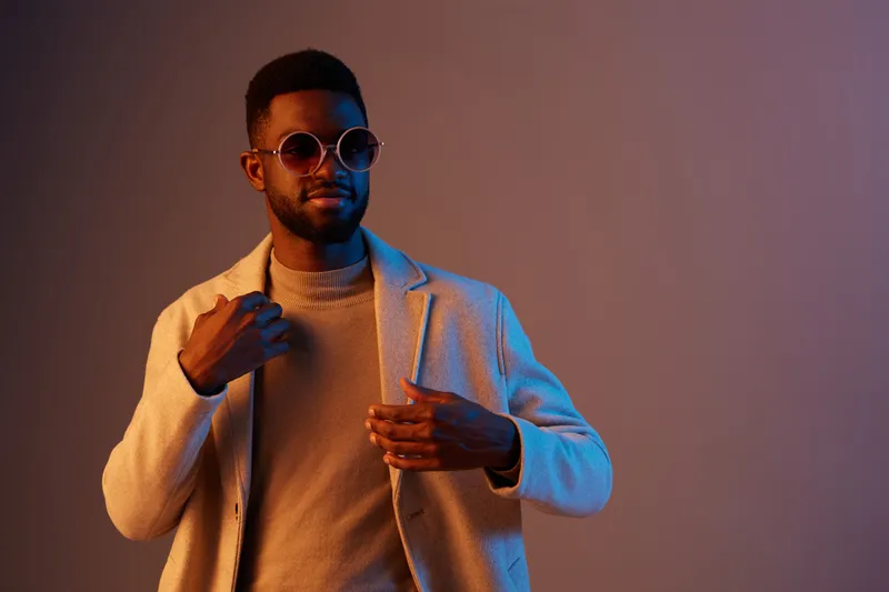 Stylish young African man in sunglasses and a fashionable coat against a vibrant gradient background, exuding confidence and modernity