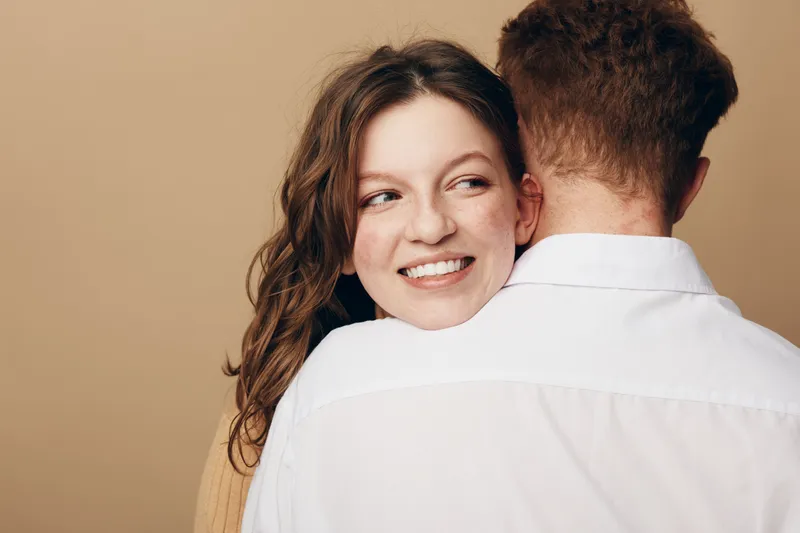 Smiling woman embraces man wearing white shirt, looking away with happy expression. Intimate moment captured in warm studio lighting on beige background, concept of love and affection.
