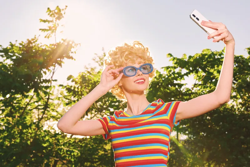 Outdoors selfie of a smiling woman in a striped colorful shirt with retro film color, wearing sunglasses, capturing a sunny park moment with cheerful vibe