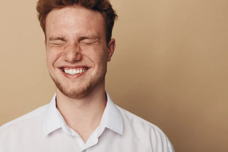 Smiling young man with red hair and freckles closes eyes in joyful expression. Casual white shirt and beige background create warm, happy, and natural portrait concept.