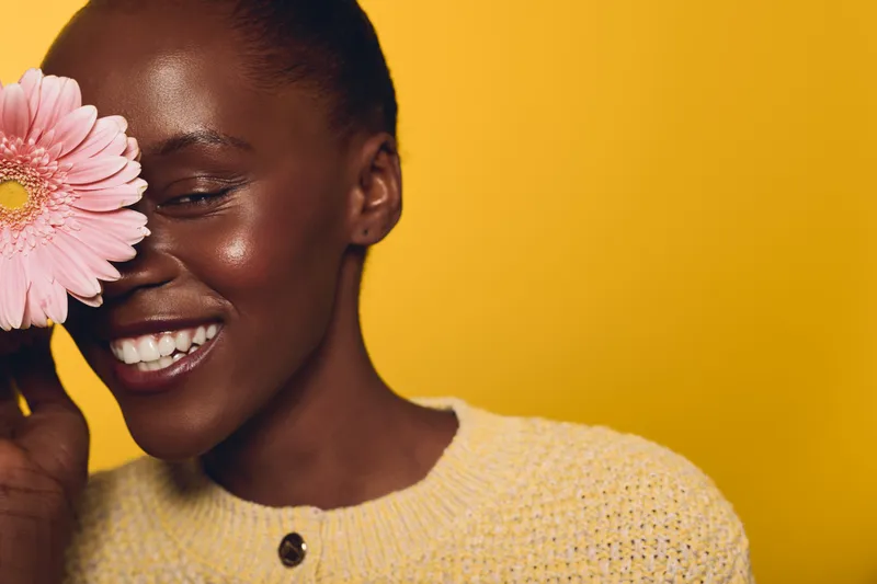 Happy African woman with dark skin smiles holding pink flower near face on yellow background. Concept of natural beauty, joy, floral, skincare and positive mood in studio portrait.