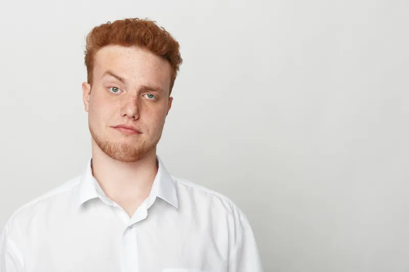 Portrait of a young man with red hair and beard wearing a white shirt against a plain light background. Casual style, confident expression, natural lighting, studio setting.