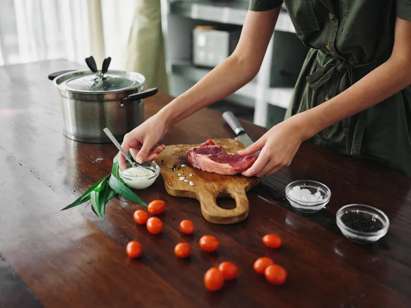 Женщина на кухне готовит ужин из мяса, овощей и специй на деревянном столе на фоне стильной кухни, крупный план рук, образ жизни