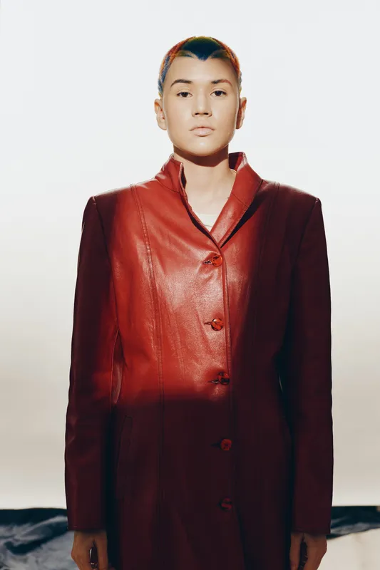 Young woman wearing a stylish red leather coat standing confidently against a minimal light background with a calm expression and modern haircut.